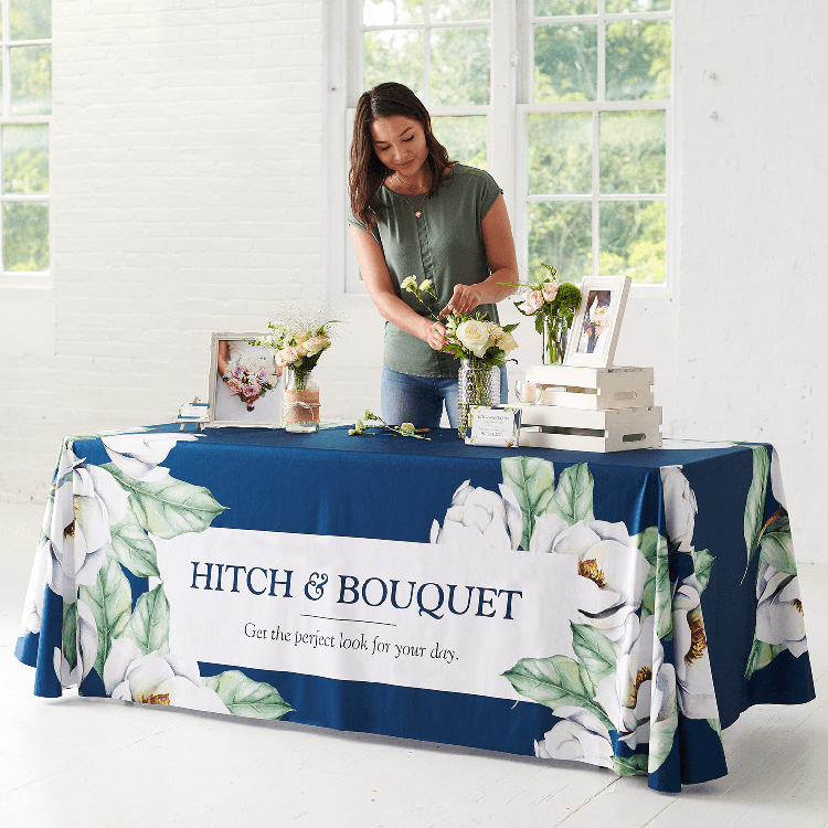 An attractive custom printed tablecloth featuring an all-over print design. A lady is standing behind it arranging some products on top of it.